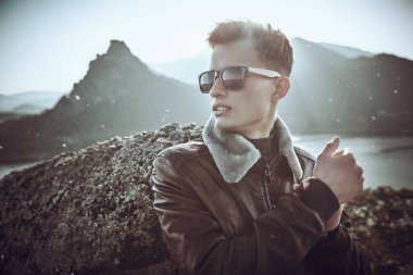 Deri ceketli ve siyah güneş gözlüklü yakışıklı genç bir adam dağın tepesinde duran doğanın güzelliğine bakıyor. Dağ manzarası arka planı. 