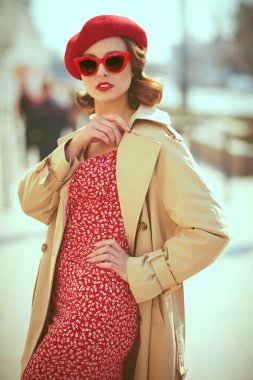 Zarif bir retro tarzı. Kırmızı elbiseli, yağmurluklu, kırmızı bereli ve zarif kırmızı gözlüklü güzel bir genç kadın bir şehir caddesinde duruyor. Güzellik, moda.