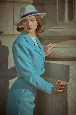 Hollywood tarzı. Zarif bir gökyüzü mavi takım elbiseli ve beyaz şapkalı güzel kahverengi saçlı bir kadın, şehrin bir caddesinde binanın bir köşesinde duruyor. 40 'lı yılların tarzı. Makyaj ve saç stili.
