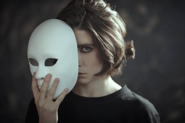 Human roles. Hypocrisy. Portrait of a brunette girl in black clothes peeking out from behind a mask-role in her hand on a dark grunge background. Mental disorders.  clipart