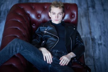 Siyah deri ceketli ve siyah kotlu şık bir genç çatı katındaki deri bir kanepede dinleniyor. Erkek modası ve tarzı.