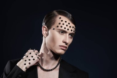 Stüdyoda, siyah ceketli yakışıklı bir erkek mankenin yüzünde ve elinde metal çivilerle poz verirken çekilmiş. Koyu arkaplan.