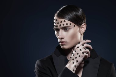 Moda fotoğrafı. Siyah kuyruklu yakışıklı, vahşi bir adamın portresi. Yüzünde ve elinde siyah bir arka planda metal çiviler var. Boşluğu kopyala.