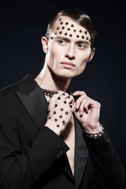 Yakışıklı, vahşi bir adam siyah bir ceketle poz veriyor. Yüzünde metal çiviler, elinde siyah bir arka plan var. Moda fotoğrafı.. 