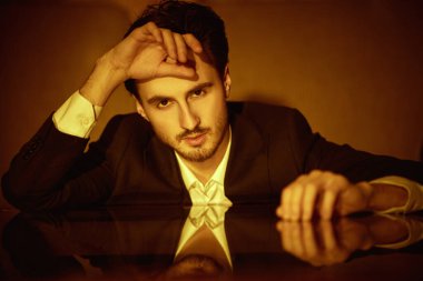 Zarif takım elbiseli yakışıklı bir esmer adam portresi ve masaya yaslanmış kameraya bakan beyaz bir gömlek. Erkek güzelliği ve modası.