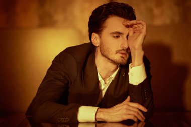 Zarif siyah takım elbiseli ve papyonlu yakışıklı bir esmer adam loş ışıklandırmalı bir odada masaya uzanıp duruyor. Erkek güzeli, moda. Kara film stili. 