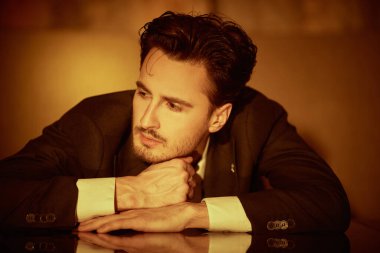 Zarif takım elbiseli, beyaz gömlekli yakışıklı bir adamın portresi masaya yaslanmış ve düşünceli bir şekilde uzağa bakıyor. Erkek güzelliği ve modası. 