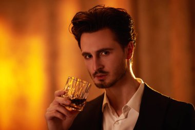 Siyah takım elbiseli, beyaz gömlekli zarif bir esmer adamın portresi loş ışıklandırmalı ve viskili bir odada duruyor. Erkek güzelliği, yaşam tarzı. Kara film stili.