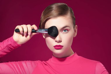 Güzel sarışın bir kız parlak pembe rujlu ve gözlerinde pembe oklar olan gözünün yanında makyaj fırçası tutan. Fuchsia geçmişi. Makyaj ve makyaj malzemeleri. Makyaj araçları.