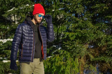 Kış kıyafetleri ve güneş gözlükleriyle yakışıklı bir adam kış parkında köknar ağaçlarının arasında yürüyor. Kış modası. Boşluğu kopyala.