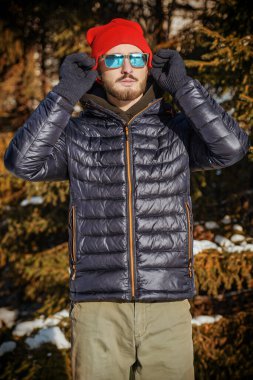 Kışlık ceketli yakışıklı bir esmer adam portresi örgü bir şapka ve güneş gözlüğü kış parkında karla kaplı köknar ağaçlarının yanında duruyordu. Kış modası.