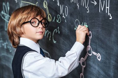 Eğitim. Temiz okul üniforması ve gözlüklü akıllı bir çocuk tahtaya oturmuş kimyasal formüller ve şemalar yazıyor. Akıllı çocuklar..  