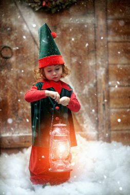 Sihirli Noel gecesi. Ellerinde yanan bir fenerle bir ahşap evin yanında duran küçük, şirin bir elf kızının uzun bir görüntüsü. Noel için mucizeler.