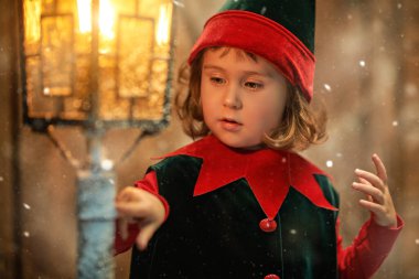 Ahşap bir kulübede duran sevimli küçük bir elf kızı kar altında karla kaplı bir fenere ilgiyle bakıyor. Kış Noel Masalı.