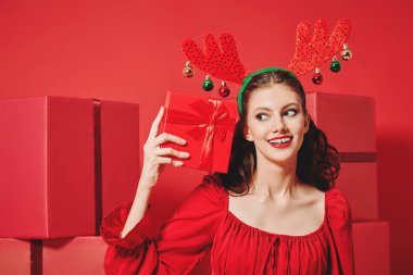 Güzel kırmızı elbiseli, akşam makyajı ve saç stili olan hoş bir kız içinde ne olduğunu düşünerek bir hediye kutusu tutuyor. Bir sürü kırmızı hediye kutusu var. Noel ve Yeni Yıl hediyeleri. 