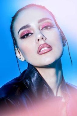 Kozmetik ve makyaj malzemeleri. Parlak gece makyajlı, siyah ceketli, mavi arka planda poz veren şehvetli esmer bir kızın portresi. Stüdyo çekimi. 