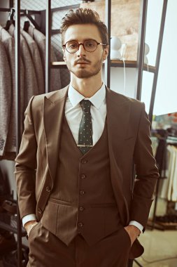 İş stili. Erkek giyim mağazasında duran kahverengi takım elbiseli esmer bir adamın portresi. Erkeklerin iş modası.