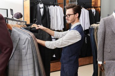 Zarif klasik takım elbiseli ve gözlüklü yakışıklı bir esmer premium erkek giyim mağazasından yeni bir takım elbise seçiyor. Erkek işi modası. Yan plan çekimi.