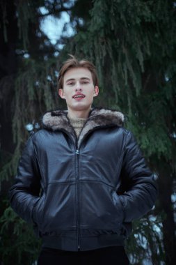 Erkekler için kış modası. Şık, siyah deri ceketli yakışıklı sarışın bir adam kış ormanında sevinçle gülümsüyor. Kış mevsimi.