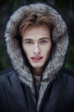 Süslü, siyah deri ceketli, başında kürk kapüşonlu çekici sarışın bir adam kış günü dışarıda dikiliyor. Erkekler kış modası.