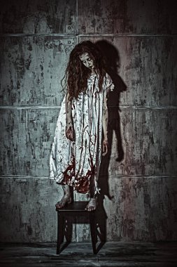 Kana bulanmış bir kadının, karanlık bir odada, bir taburede durduğu korkunç bir sahne. Kana susamış zombi kadın. Korku filmi, gerilim. Cadılar Bayramı.