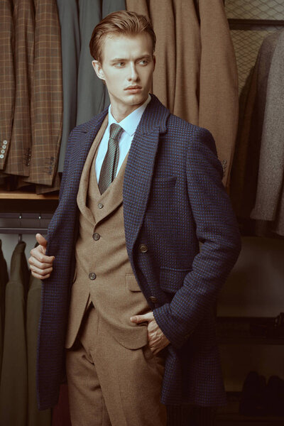 Портрет серьезного привлекательного делового человека, одетого в элегантный костюм из трех частей и пальто, стоящего на фоне роскошных классических костюмов в магазине. Мужская мода и стиль.