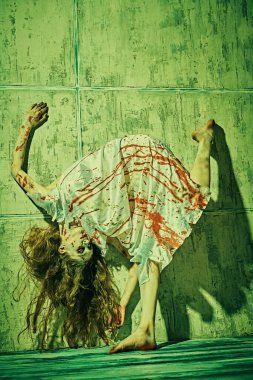 Portrait of a creepy, devil-possessed woman with pale eyes and tousled hair wearing  bloody nightie slides down the blood-covered wall. Zombie woman. Halloween.  clipart