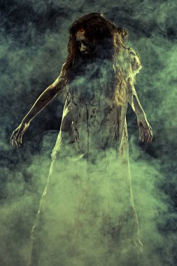 Korkunç şekilsiz bir kadın, şeytan tarafından ele geçirilmiş, kanlı bir gecelikle karanlıkta duruyor, dumanla çevrili. Ürkütücü zombi kadın. Korku, gerilim. Cadılar Bayramı.