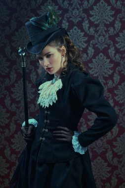 19. yüzyıl moda ve güzelliği. Zarif, siyah 19. yüzyıl takım elbiseli ve şapkalı genç bir bayan elinde bastonuyla klasik bir arka planda poz veriyor.. 