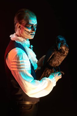 Siyah arka planda gizemli karanlık ışıklı siyah bir kuzgun pozu veren zarif elbiseli korkunç iskelet adam. Dia de los muertos. Ölüler Günü. Cadılar Bayramı. 