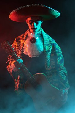 Ulusal kostümlü Meksikalı sanatçı ve Kafatası şekeri makyajlı Ölü Karnavalı 'nda gitar çalıyor. Mistik sisli ve kırmızı ışıklı siyah bir arka plan. Cadılar Bayramı.  