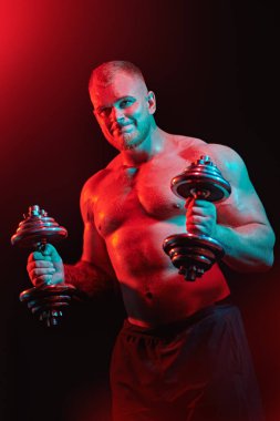 Güçlü, atletik bir adam kırmızı ve yeşil ışıkta siyah arka planda dambıllarla sıkı çalışıyor. Erkek güzellik konsepti. Fitness, vücut geliştirme.