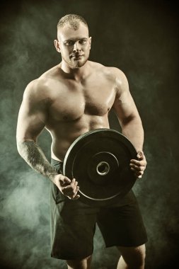 Spor konsepti, halter, vücut geliştirme. Elinde demir bir disk tutan kaslı, yakışıklı bir atletin portresi. Dumanlı siyah arkaplan.