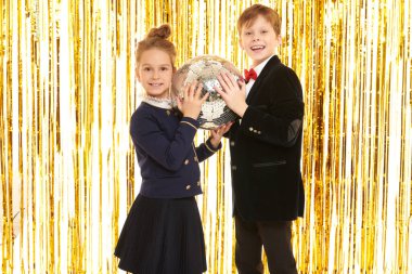 Çocuk partisi. Gece kıyafetleri içinde neşeli küçük bir kız ve oğlan, arka plandaki parlak altın folyo perdelerde bir disko topuyla neşeli bir partide poz veriyorlar. Çocuk modası ve tatiller. 