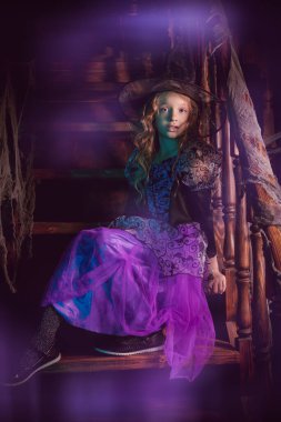 Mor cadı kostümlü şirin küçük kız merdivenlerde ağla kaplı oturuyor. Eski evin karanlık ve gizemli bir yerinde tam boy portre. Cadılar Bayramı.