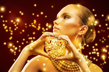 Gorgeous young woman with perfect skin and golden makeup and hair holds heart shaped christmas toy in her hands. Dark red background with shining golden lights. Gold Christmas luxury style.  clipart