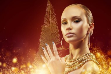 Lüks şenlik tarzı. Altın tenli, makyajlı altın takılar takan ve elinde altın tüy olan güzel bir kız. Sihirli altın ışıkları olan koyu kırmızı bir arka plan. Moda altın mücevheri.
