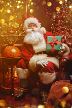 Geleneksel Noel ve Yeni Yıl. Nazik Noel Baba elinde hediyesiyle güzel bir Noel ortamında bir koltukta oturur. Noel ışıkları parlıyor.