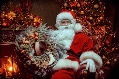 Geleneksel Noel ve Yeni Yıl. Nazik Noel Baba elinde Noel çelengiyle güzel bir Noel ortamında bir sandalyede oturuyor ve gülümsüyor.. 