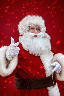 Komik ve neşeli bir Noel Baba 'nın portresi. Gözlerini kırpıyor ve parmağını kameraya doğrultuyor. Yıldızlı şenlikli kırmızı stüdyo arka planı. Noel ve Yeni Yıl.