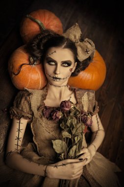 Cadılar Bayramı. Voodoo masalları. Vudu bebeği gibi giyinmiş güzel ve tehlikeli genç bir kadın ahşap bir zeminde uzanmış, balkabaklarına yaslanmış ve ellerinde kurumuş güller tutuyor..