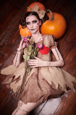 Voodoo tales. A beautiful and dangerous young woman dressed as a voodoo doll lies on a wooden floor, leaning on pumpkins and holding dried roses in her hands. Halloween. clipart