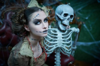 Voodoo büyüsü. Esrarengiz ve korkutucu bir kızın portresi, voodoo bebeği gibi giyinmiş, eski bir koltukta iskeletle oturuyor. Örümcek ağlarıyla kaplı kasvetli eski bir ev. Cadılar Bayramı.