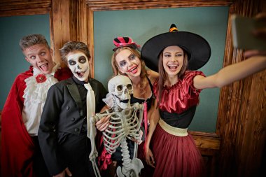 Merry Halloween party. Group of cheerful friends teenagers make selfie in the scenery of an old fairy-tale castle. Vampire, clown, witch and skeleton. clipart