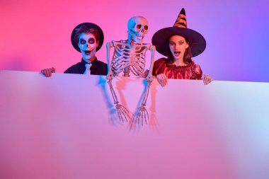 Güzel genç cadı ve şeker kafalı genç bir çocuk iskelet ve fotokopi alanı olan bir tabela tutuyorlar. Cadılar Bayramı kutlaması. Kostümler, makyaj ve saç stili. Renkli ışıklandırmalı stüdyo portresi.