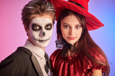 Güzel, genç bir cadı ve iskelet gibi genç bir oğlan. Cadılar Bayramı kutlaması. Cadılar Bayramı için kostüm, makyaj ve saç stili. Renkli ışıklandırmalı stüdyo portresi.