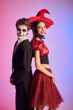 Güzel, genç bir cadı ve iskelet gibi genç bir oğlan. Cadılar Bayramı kutlaması. Cadılar Bayramı için kostüm, makyaj ve saç stili. Renkli ışıklandırmalı stüdyo portresi.
