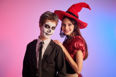 Güzel, genç bir cadı ve iskelet gibi genç bir oğlan. Cadılar Bayramı kutlaması. Cadılar Bayramı için kostüm, makyaj ve saç stili. Renkli ışıklandırmalı stüdyo portresi.