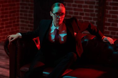 Şahane kendine güvenen bayan şık bir takım elbiseyle deri koltukta kravat ve gözlüklerle poz veriyor. Lüks bir iş tarzı. Gözlük modası. Karışık ışıklandırma. 