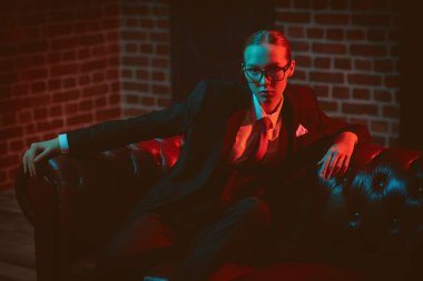 Şahane kendine güvenen bayan şık bir takım elbiseyle deri koltukta kravat ve gözlüklerle poz veriyor. Lüks bir iş tarzı. Gözlük modası. Karışık ışıklandırma. 